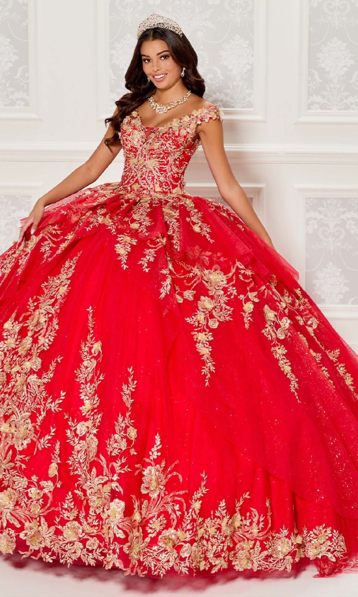 A young woman in a jeweled tiara and necklace wears the Quinceanera Dress PR30119 By Princesa, featuring off-the-shoulder gold floral embroidery and glitter tulle, standing before an ornate white wall.
